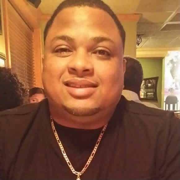 A smiling man wearing a black shirt and a chain necklace, seated in a restaurant. — Deputy killed
