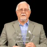 Curtis Brewer holding a Hall of Fame plaque, wearing a suit and glasses, with a serious expression.