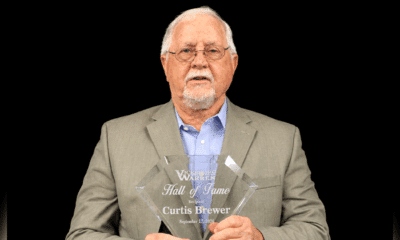 Curtis Brewer holding a Hall of Fame plaque, wearing a suit and glasses, with a serious expression.