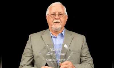 Curtis Brewer holding a Hall of Fame plaque, wearing a suit and glasses, with a serious expression.
