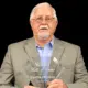 Curtis Brewer holding a Hall of Fame plaque, wearing a suit and glasses, with a serious expression.