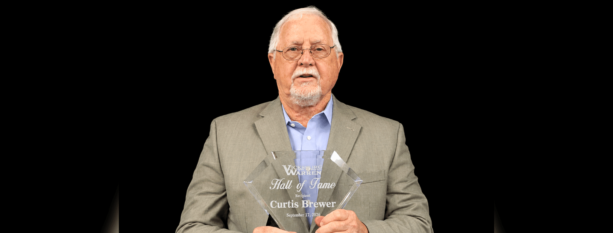 Curtis Brewer holding a Hall of Fame plaque, wearing a suit and glasses, with a serious expression.