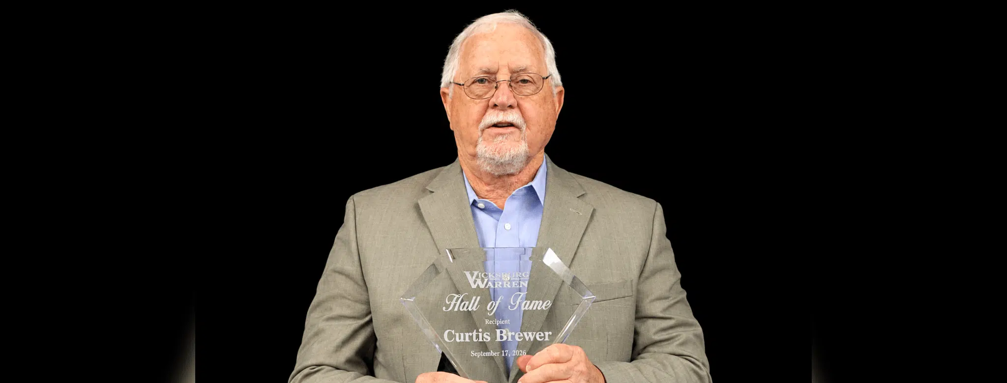 Curtis Brewer holding a Hall of Fame plaque, wearing a suit and glasses, with a serious expression.