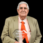 William Cook proudly holds his Hall of Fame plaque, wearing a suit and orange tie against a black background.
