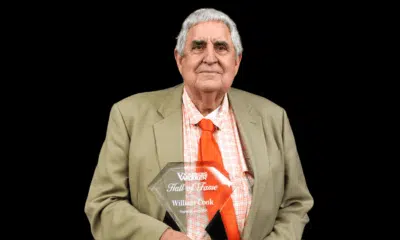 William Cook proudly holds his Hall of Fame plaque, wearing a suit and orange tie against a black background.