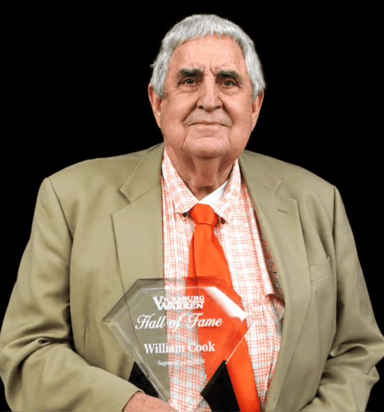 William Cook proudly holds his Hall of Fame plaque, wearing a suit and orange tie against a black background.