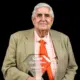 William Cook proudly holds his Hall of Fame plaque, wearing a suit and orange tie against a black background.