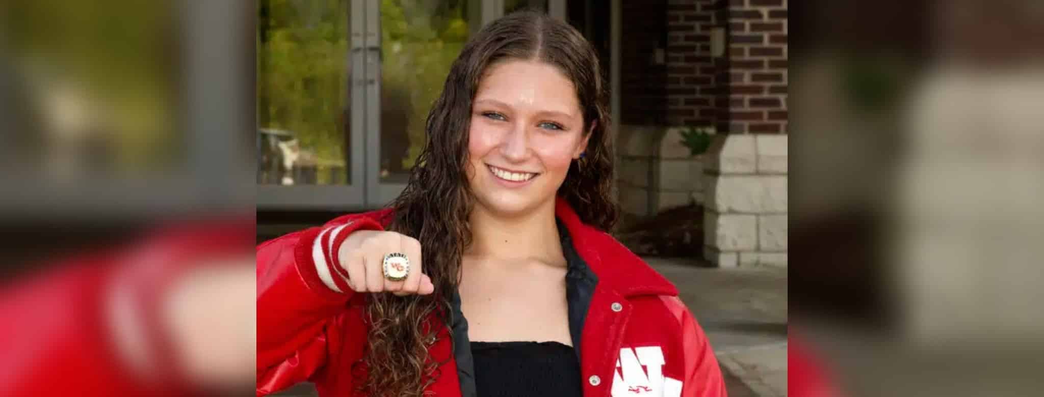 A swimmer in a red jacket proudly displays a championship ring, smiling at the camera. — Splash 4 Gold Meet