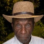 An elderly man wearing a straw hat and white shirt, with a serious expression, surrounded by greenery. — Leon Jefferson