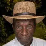An elderly man wearing a straw hat and white shirt, with a serious expression, surrounded by greenery. — Leon Jefferson