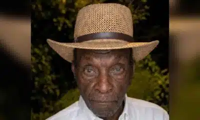 An elderly man wearing a straw hat and white shirt, with a serious expression, surrounded by greenery. — Leon Jefferson