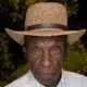 An elderly man wearing a straw hat and white shirt, with a serious expression, surrounded by greenery. — Leon Jefferson