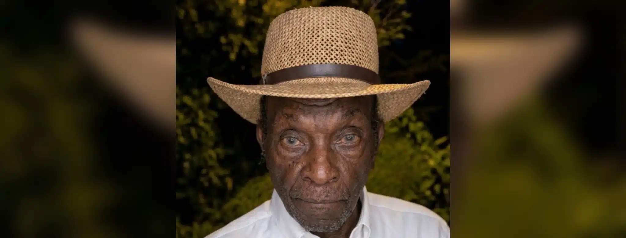 An elderly man wearing a straw hat and white shirt, with a serious expression, surrounded by greenery. — Leon Jefferson
