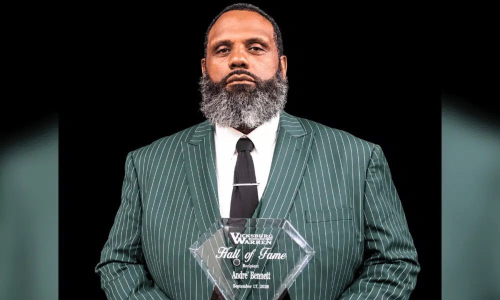 Andre Bennett holding a Hall of Fame plaque, wearing a green striped suit and tie, with a serious expression.