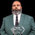 Andre Bennett holding a Hall of Fame plaque, wearing a green striped suit and tie, with a serious expression.