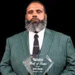Andre Bennett holding a Hall of Fame plaque, wearing a green striped suit and tie, with a serious expression.