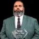 Andre Bennett holding a Hall of Fame plaque, wearing a green striped suit and tie, with a serious expression.