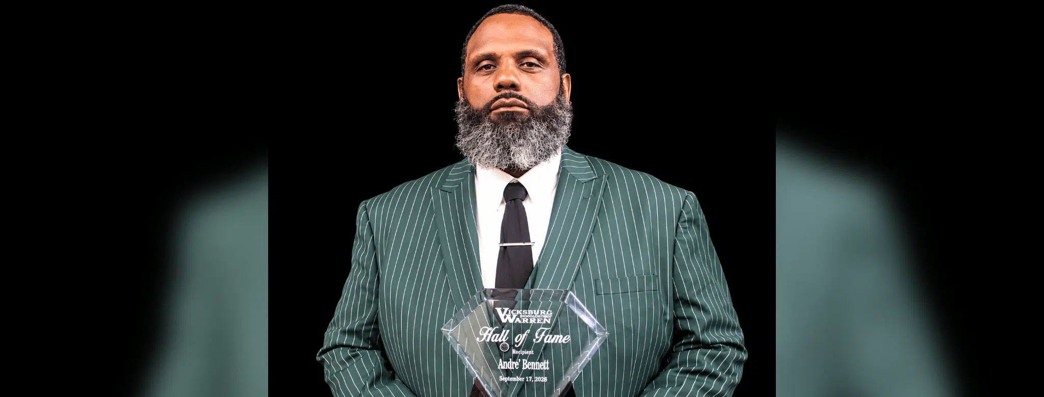 Andre Bennett holding a Hall of Fame plaque, wearing a green striped suit and tie, with a serious expression.