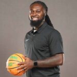 DeAndre King, a basketball coach, smiles while holding a basketball in a black polo shirt.
