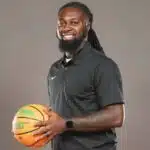DeAndre King, a basketball coach, smiles while holding a basketball in a black polo shirt.