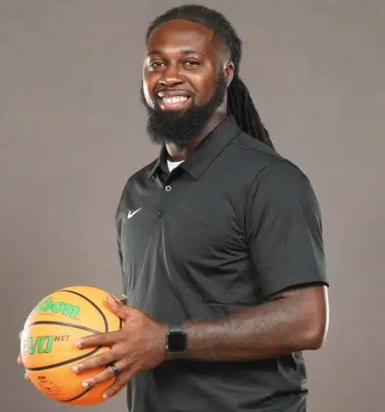 DeAndre King, a basketball coach, smiles while holding a basketball in a black polo shirt.