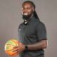 DeAndre King, a basketball coach, smiles while holding a basketball in a black polo shirt.