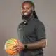 DeAndre King, a basketball coach, smiles while holding a basketball in a black polo shirt.