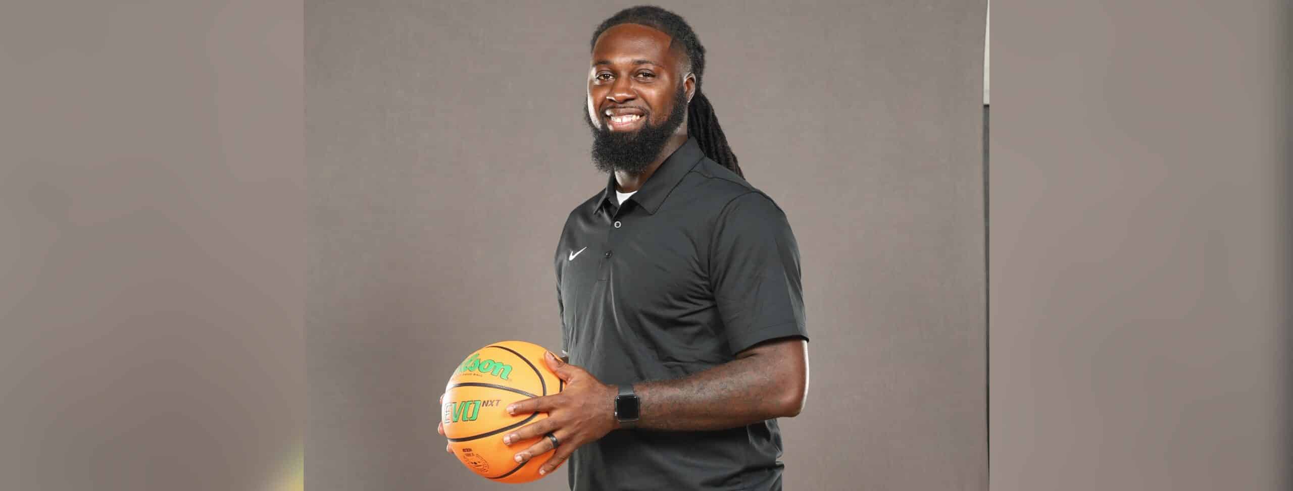 DeAndre King, a basketball coach, smiles while holding a basketball in a black polo shirt.