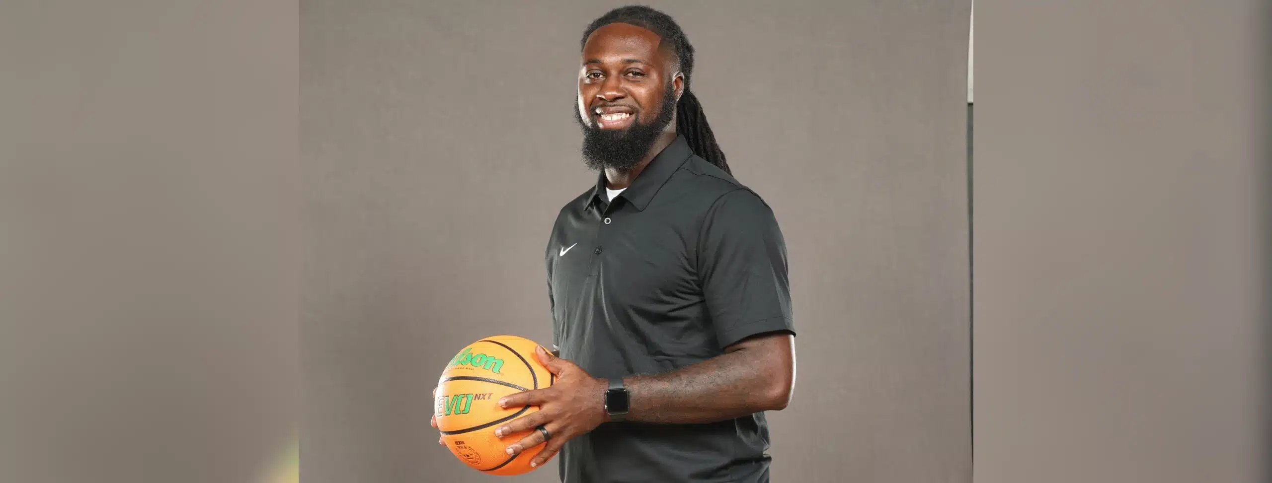 DeAndre King, a basketball coach, smiles while holding a basketball in a black polo shirt.