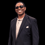 Smiling man in sunglasses and a black suit, standing confidently against a dark background. — Cleothia Williams