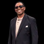 Smiling man in sunglasses and a black suit, standing confidently against a dark background. — Cleothia Williams