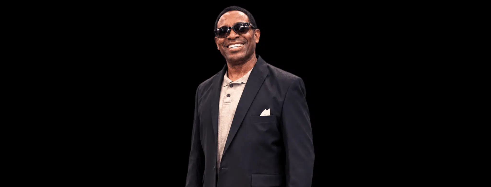Smiling man in sunglasses and a black suit, standing confidently against a dark background. — Cleothia Williams