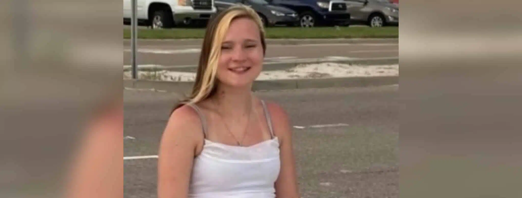 A young woman with long blonde hair smiles while sitting outdoors, wearing a white tank top. — Avery Read Carroll