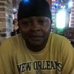 A man wearing a yellow New Orleans Saints t-shirt sits indoors, smiling softly. — Alan Fredrick Lyons jr