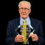 Jim Taylor holding his Hall of Fame plaque, wearing a suit and tie, with a serious expression.