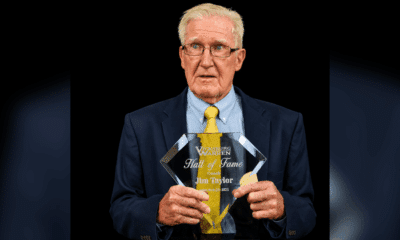 Jim Taylor holding his Hall of Fame plaque, wearing a suit and tie, with a serious expression.