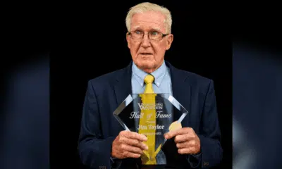 Jim Taylor holding his Hall of Fame plaque, wearing a suit and tie, with a serious expression.