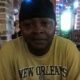 A man wearing a yellow New Orleans Saints t-shirt sits indoors, smiling softly. — Alan Fredrick Lyons jr