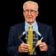 Jim Taylor holding his Hall of Fame plaque, wearing a suit and tie, with a serious expression.