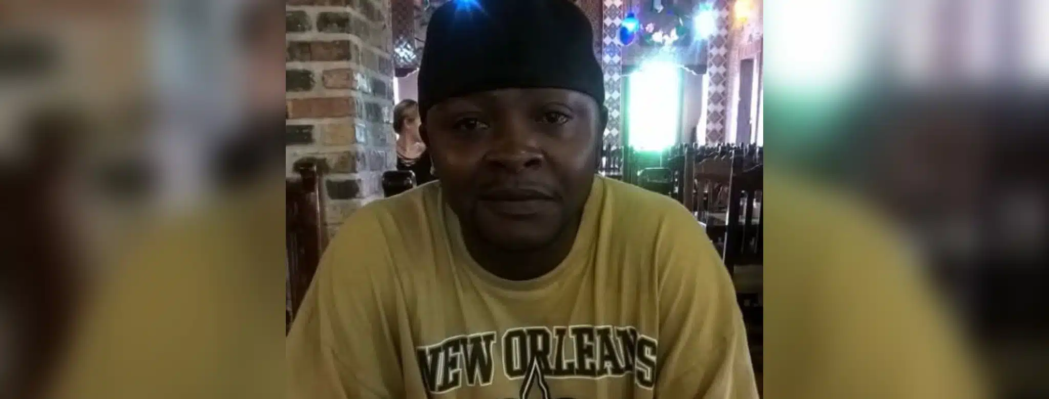 A man wearing a yellow New Orleans Saints t-shirt sits indoors, smiling softly. — Alan Fredrick Lyons jr