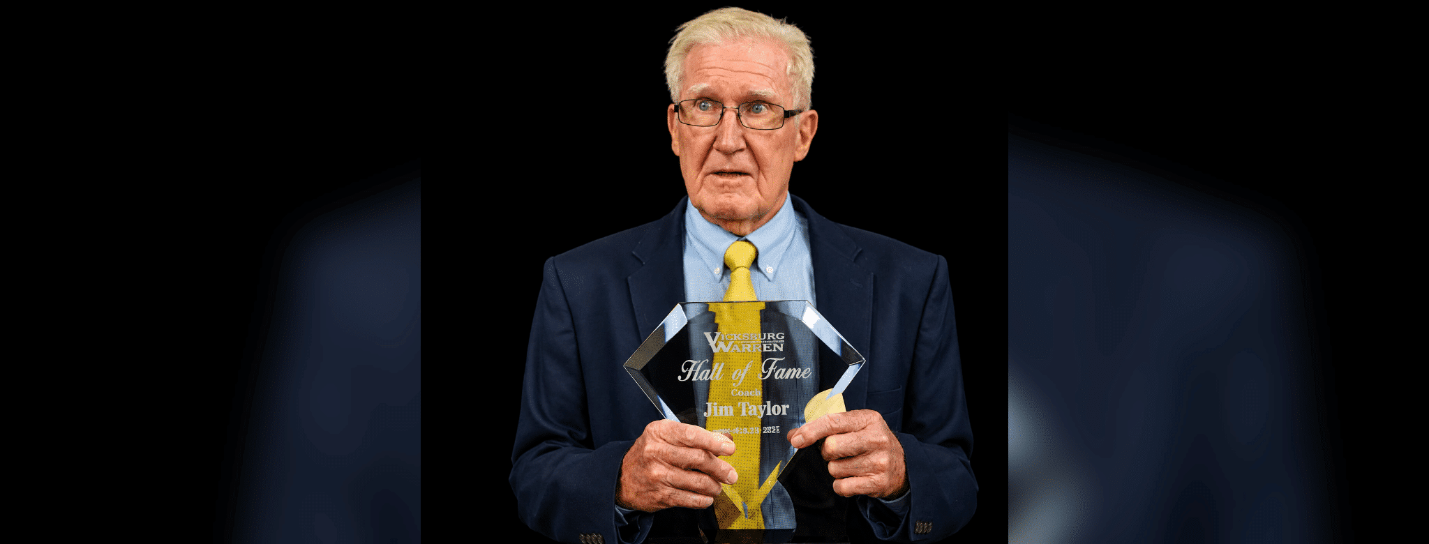 Jim Taylor holding his Hall of Fame plaque, wearing a suit and tie, with a serious expression.
