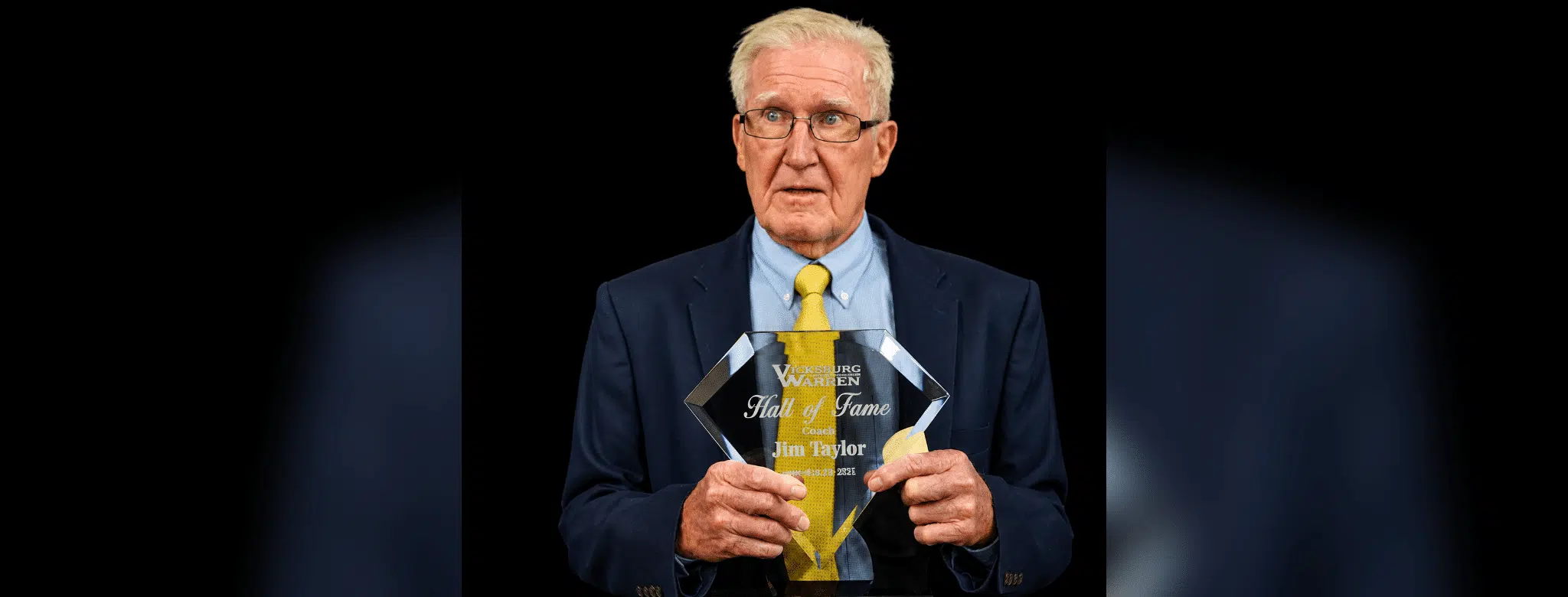 Jim Taylor holding his Hall of Fame plaque, wearing a suit and tie, with a serious expression.