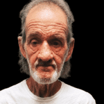 A 70-year-old man with gray hair and a beard, wearing a white shirt, looks directly at the camera against a dark background.