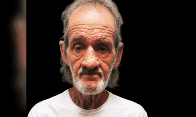 A 70-year-old man with gray hair and a beard, wearing a white shirt, looks directly at the camera against a dark background.
