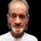 A 70-year-old man with gray hair and a beard, wearing a white shirt, looks directly at the camera against a dark background.
