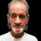 A 70-year-old man with gray hair and a beard, wearing a white shirt, looks directly at the camera against a dark background.