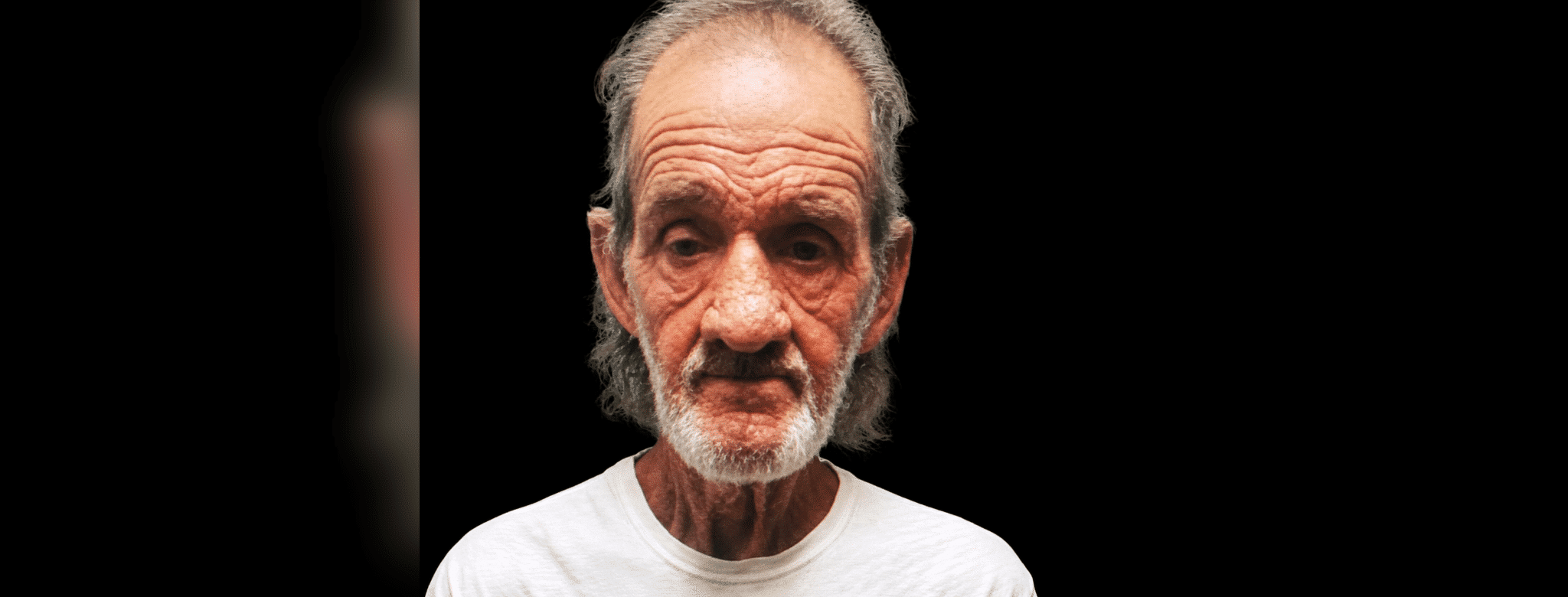 A 70-year-old man with gray hair and a beard, wearing a white shirt, looks directly at the camera against a dark background.