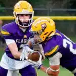 Two football players in purple and yellow uniforms executing a handoff during a — Flashes fall to Riverfield in close game