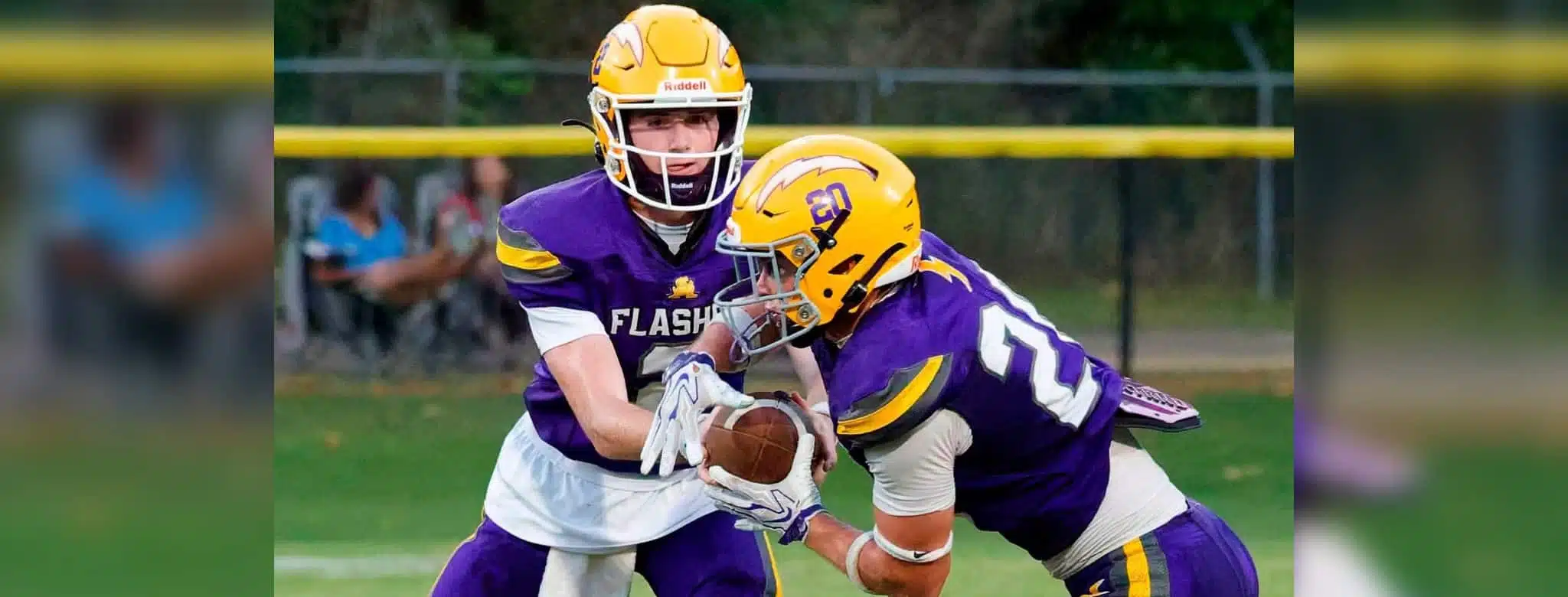 Two football players in purple and yellow uniforms executing a handoff during a — Flashes fall to Riverfield in close game