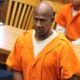 A man in an orange prison jumpsuit stands in a courtroom, looking serious, with other individuals se — Failure to register